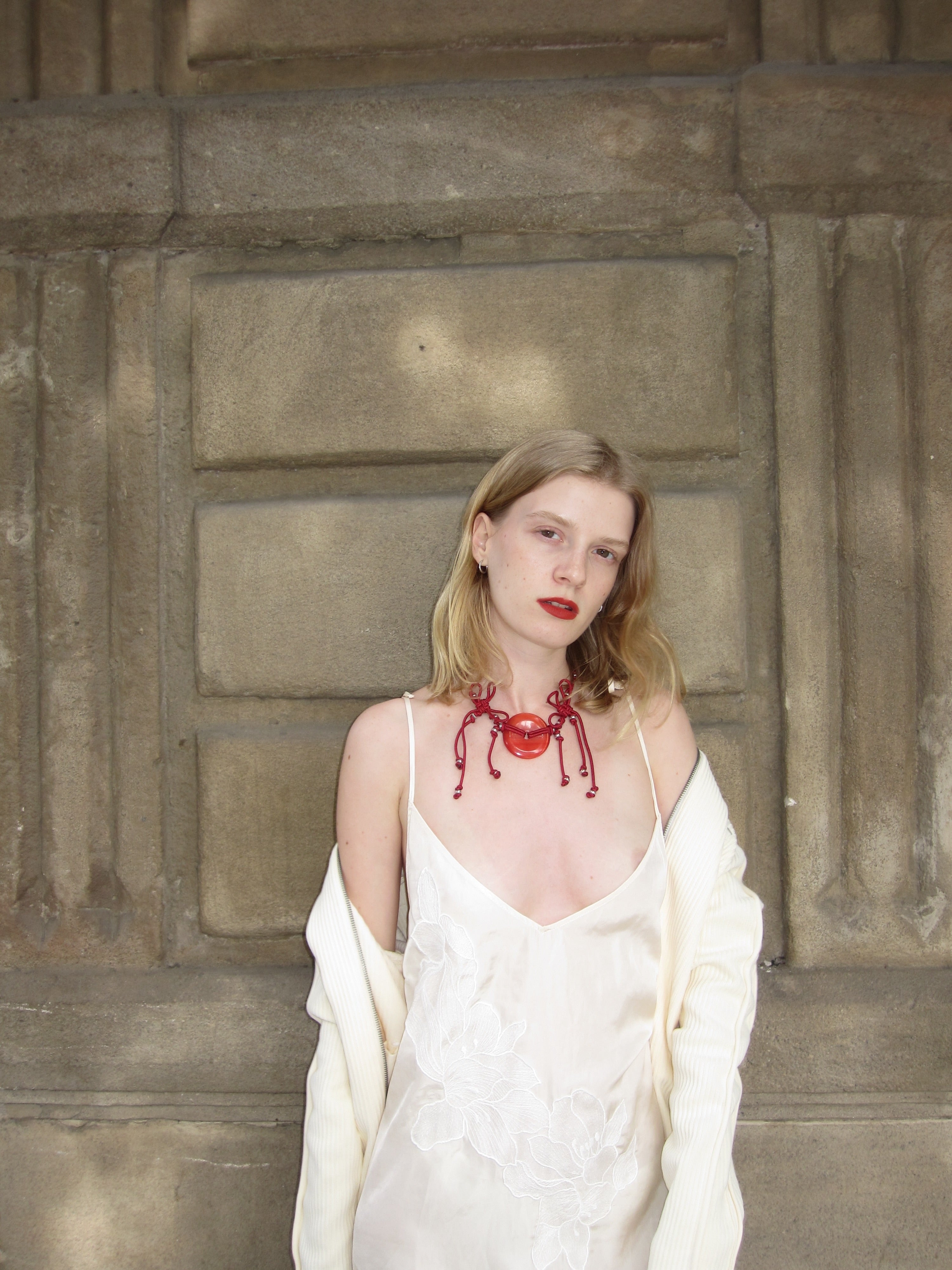 Woman wearing a white dress with a red necklace against a textured wall.