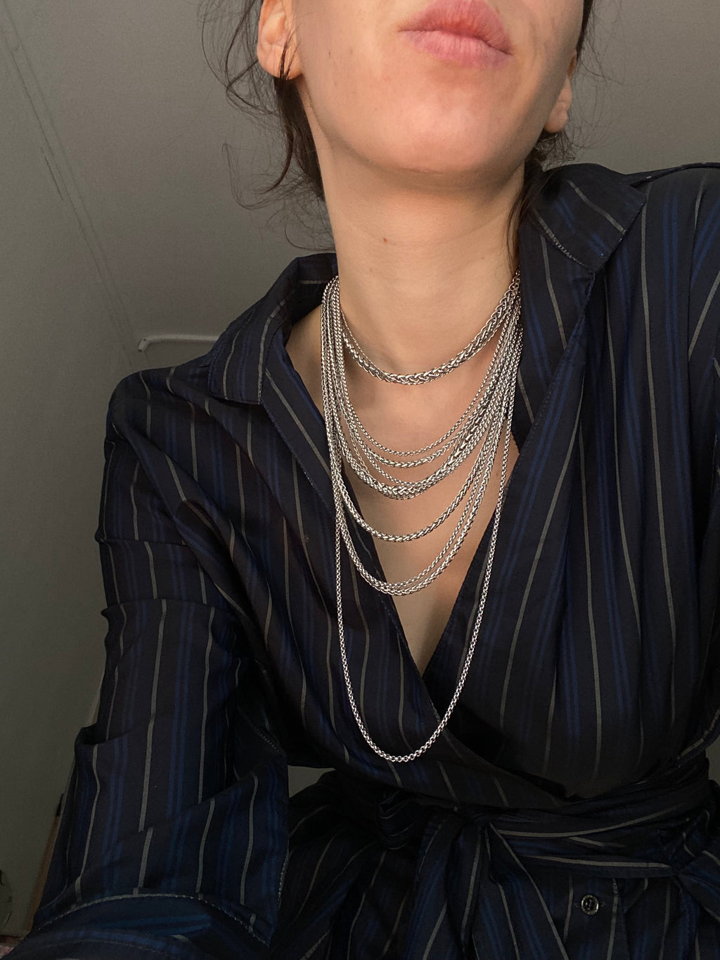 Person wearing multiple silver necklaces with a dark striped shirt against a neutral background