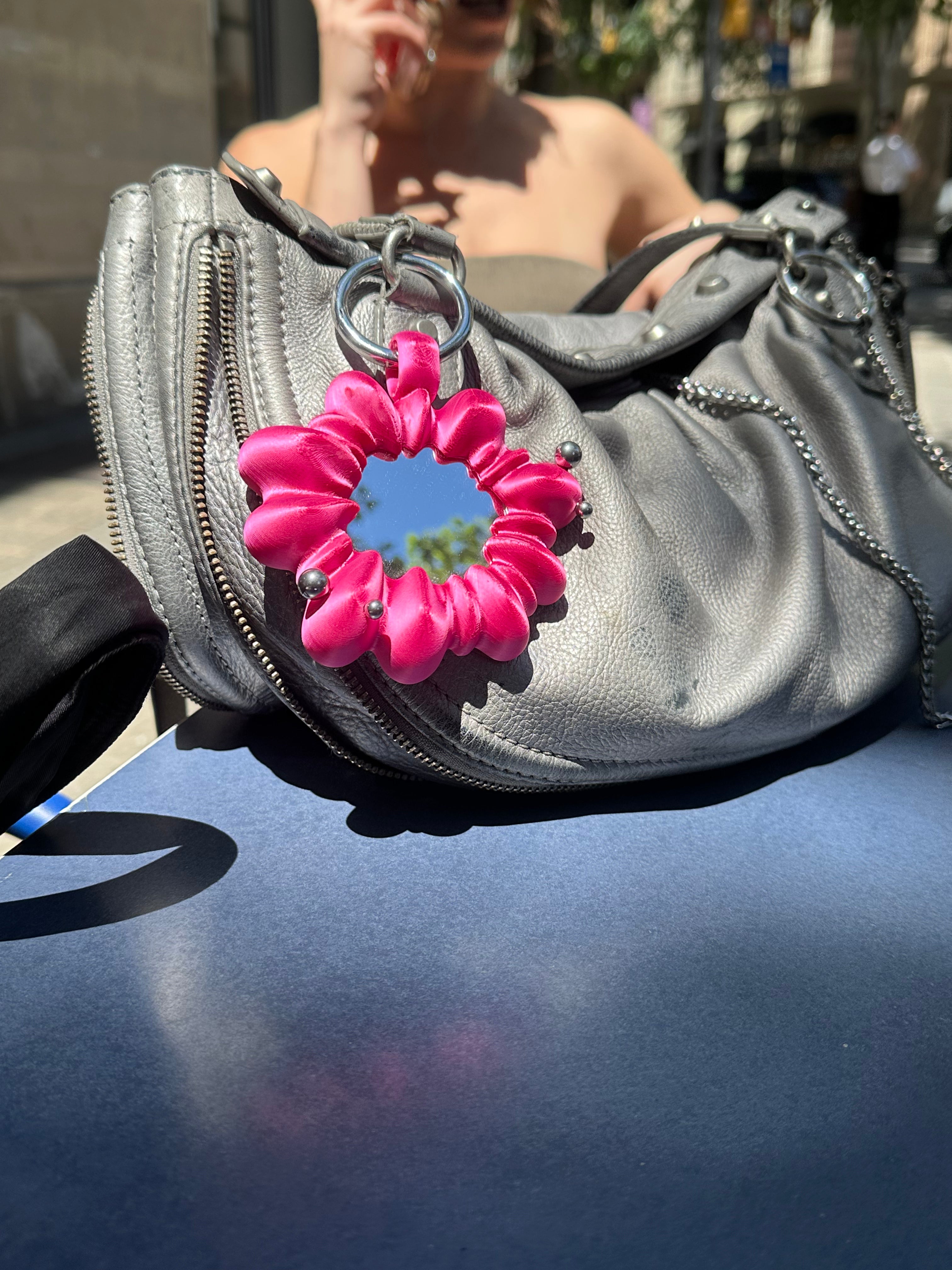 Gray handbag with a pink flower-shaped mirror attached on a reflective surface.