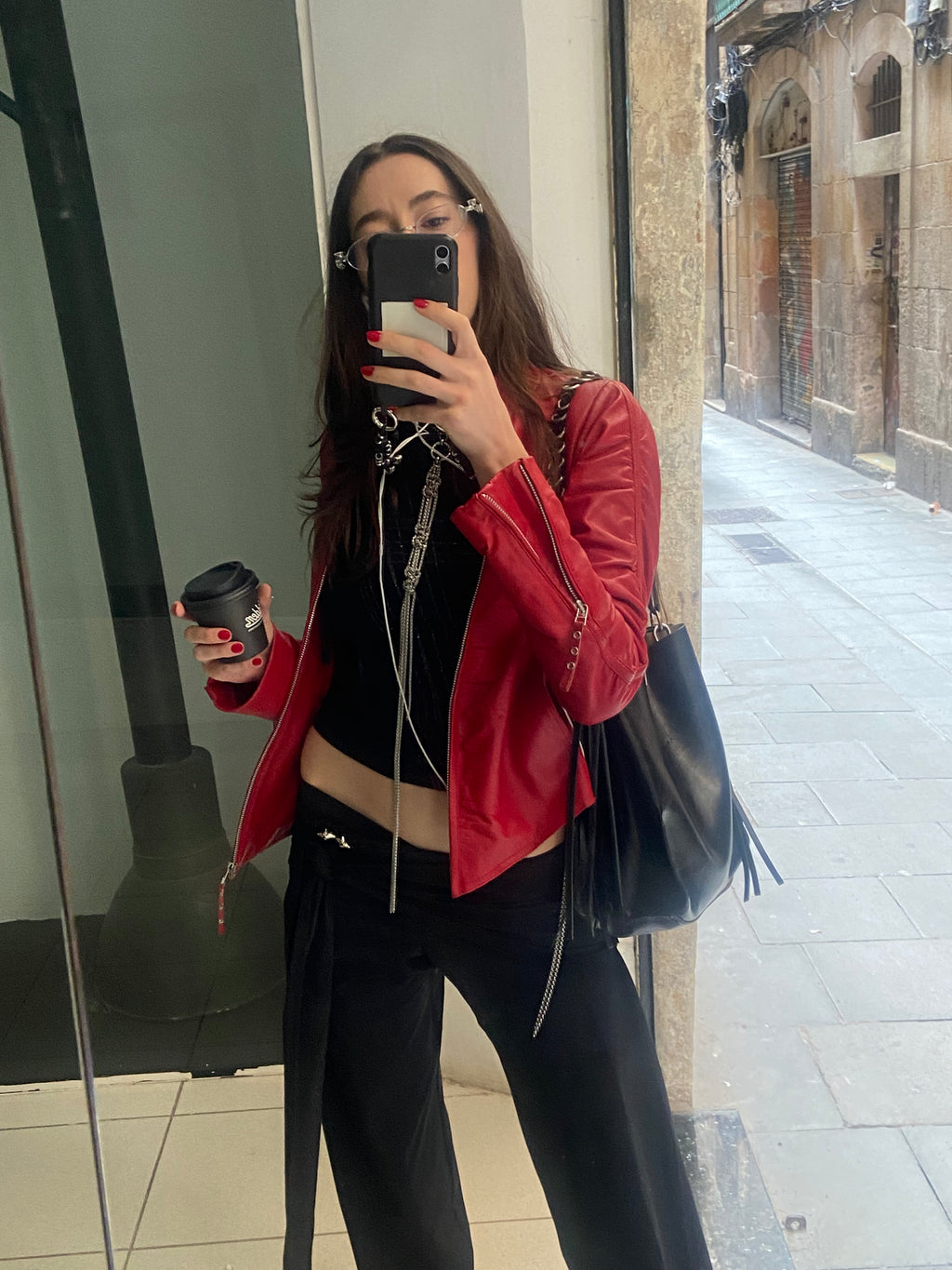 Woman in a red jacket holding a coffee cup and phone, standing in front of a mirror.